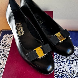 May include: A pair of black leather pumps featuring a rounded toe, a low heel, and a black grosgrain bow on the vamp accented with a gold-tone metal plate engraved with the Salvatore Ferragamo logo. The interior insole is stamped with the brand name.
