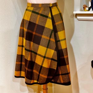 Pode incluir: Saia vintage envelope com padrão xadrez amarelo, castanho e verde. A saia tem um fecho de botão na cintura e uma barra preta ao longo da bainha. A saia é exibida em um manequim de madeira.