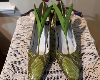 May include: A pair of green snakeskin-patterned heels with pink and white tulips. The shoes are displayed on a cream-colored, open-weave doily. The heels have a pointed toe and a low heel.