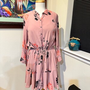 May include: A pink floral dress with long sleeves and a button-up front. The dress has a cinched waist and a tiered skirt.