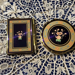 May include: Two vintage compact mirrors with gold trim and a blue background. The design features a teapot with pink flowers and gold accents.