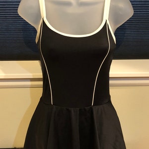 May include: A black swimsuit with white trim. The swimsuit has a scoop neck and thin straps. The skirt portion of the swimsuit is gathered and has a slight flare.