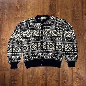 Könnte beinhalten: Schwarze und cremefarbene Strickjacke mit detailliertem geometrischem Muster. Der Pullover hat einen runden Ausschnitt, lange Ärmel und silberfarbene Knöpfe. Das Design umfasst Herz- und Diamantformen. Maße in cm.