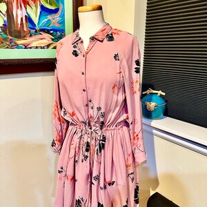 May include: A pink floral print dress with long sleeves and a button-up front. The dress has a cinched waist and a ruffled skirt.