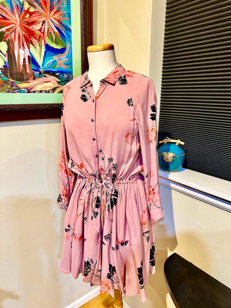 May include: A pink floral print dress with long sleeves and a button-up front. The dress has a cinched waist and a ruffled skirt.