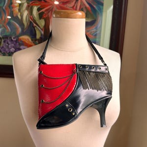 Op de afbeelding: Een unieke ketting met een rood lakleren paneel met gouden kettingaccenten, gecombineerd met een zwart ontwerp van een schoen met hoge hakken. De ketting wordt op een mannequin gepresenteerd, wat het ongewone ontwerp benadrukt. Afmetingen in cm.