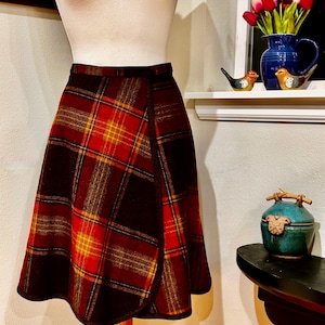Pode incluir: Uma saia envelope vintage com um padrão xadrez. A saia apresenta uma paleta de cores vermelha, castanha e amarela. A saia é exibida em um manequim. A saia tem uma silhueta clássica em forma de A.