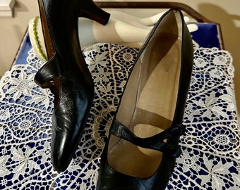 May include: A pair of black leather heeled shoes with a strap and buckle detail. The shoes are on a white lace doily.