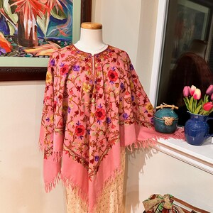 May include: A pink shawl with detailed floral embroidery in red, purple, yellow, and green. The shawl features a fringed edge and is displayed on a mannequin. The garment is part of a collection of shawls and wraps.