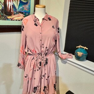 May include: A pink floral button-down dress with long sleeves and a cinched waist. The dress has a tiered skirt and a black floral print.