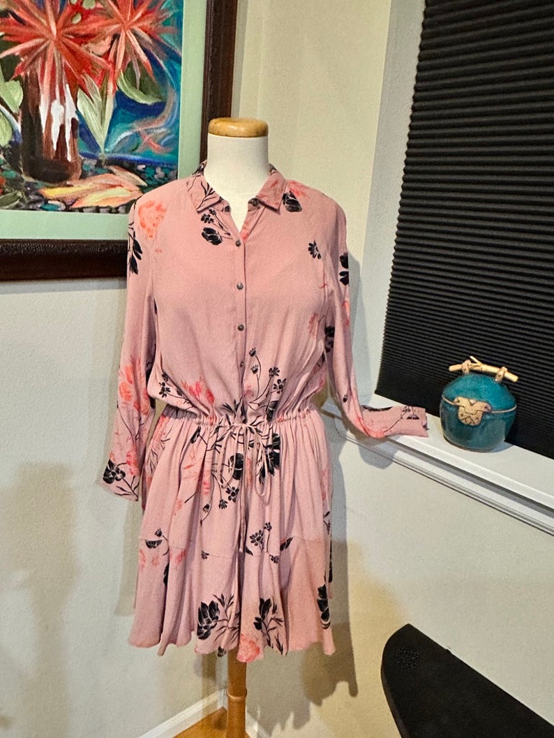 May include: A pink floral button-down dress with long sleeves and a cinched waist. The dress has a tiered skirt and a black floral print.