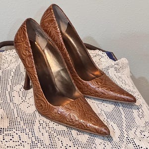 Könnte beinhalten: Ein Paar braune Leder-High Heels mit spitzer Zehenpartie. Die Schuhe haben ein geprägtes Blumenmuster und einen Stiletto-Absatz. Die Absätze werden auf einer weißen Spitzendecke präsentiert.