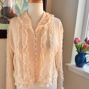 May include: A peach-colored blouse with long sleeves and a ruffled collar and trim. The blouse features a series of small pearl buttons down the front and delicate lace detailing. The fabric has a subtle polka dot pattern.
