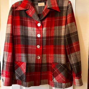 Puede incluir: Una chaqueta de manga larga con un patrón de cuadros en rojo, gris y negro. La chaqueta tiene un cuello puntiagudo, cuatro botones blancos grandes en la parte delantera central y dos bolsillos cuadrados en la parte inferior. Se muestra en una percha negra.