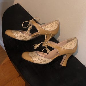 May include: A pair of tan and cream-colored vintage-style heels. The shoes feature a floral pattern on the toe and heel, with a strap and tassel detail. The heels are a curved shape.
