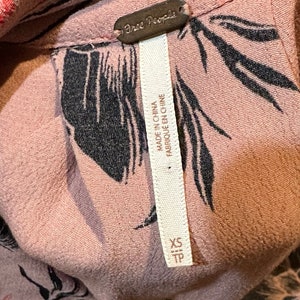 May include: A pink floral print shirt with a black Free People tag. The tag says "Made in China, Fabrique en Chine". The shirt is size XS/TP.