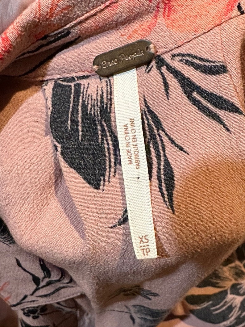 May include: A pink floral print shirt with a black Free People tag. The tag says "Made in China, Fabrique en Chine". The shirt is size XS/TP.