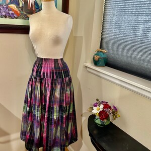 Può includere: Una gonna a quadri colorata con vita alta e design plissettato. La gonna è realizzata in tessuto leggero e ha una combinazione di colori nero, rosa, verde e viola.