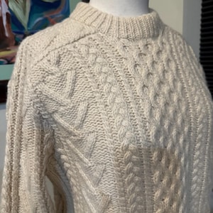 Op de afbeelding: Een crèmekleurige kabelgebreide trui met een klassieke ronde hals. De trui is gemaakt van een zacht, warm materiaal en heeft een gedetailleerd kabelgebreid patroon.