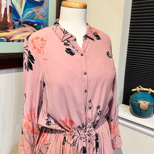 May include: A long-sleeved pink button-down dress with a floral print in black and orange. The dress has a drawstring waist and a relaxed fit.