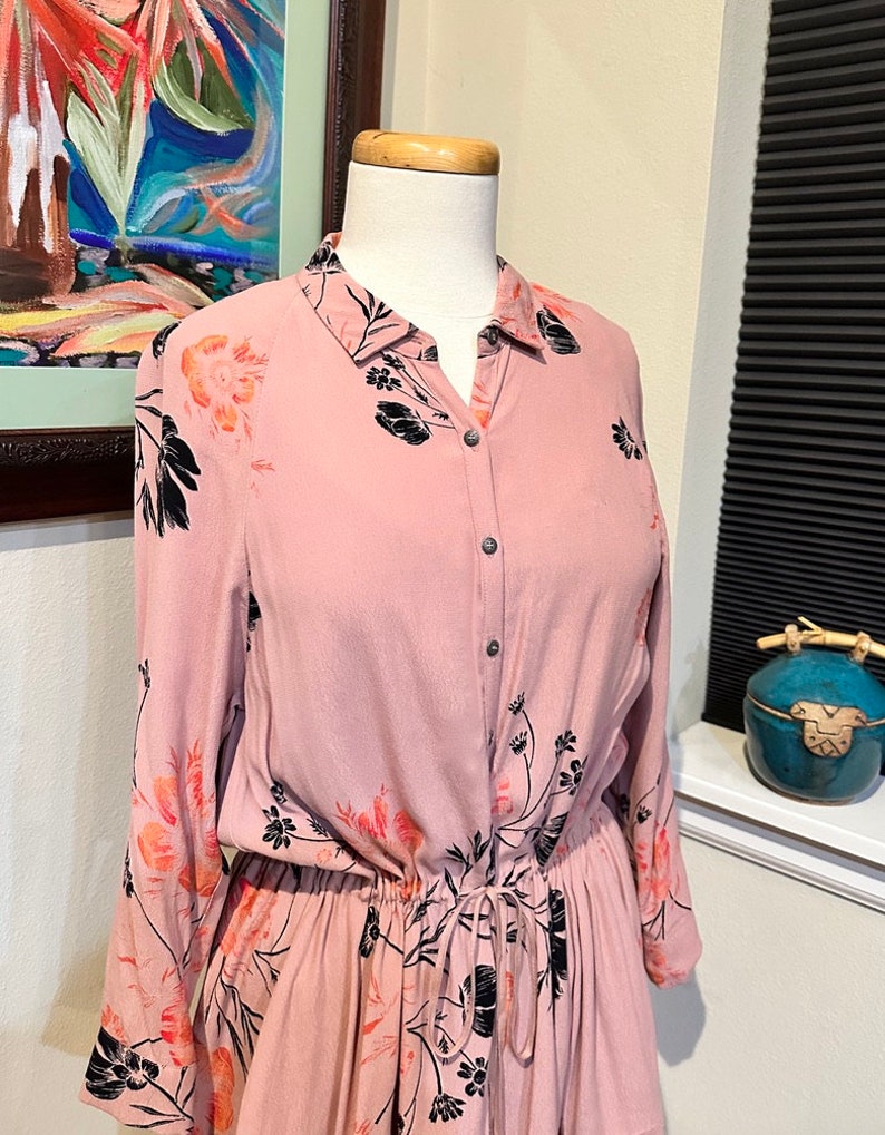 May include: A long-sleeved pink button-down dress with a floral print in black and orange. The dress has a drawstring waist and a relaxed fit.