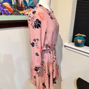 May include: A pink floral dress with black and orange floral print. The dress has long sleeves and a button-up front. The dress has a cinched waist and a ruffled hem.