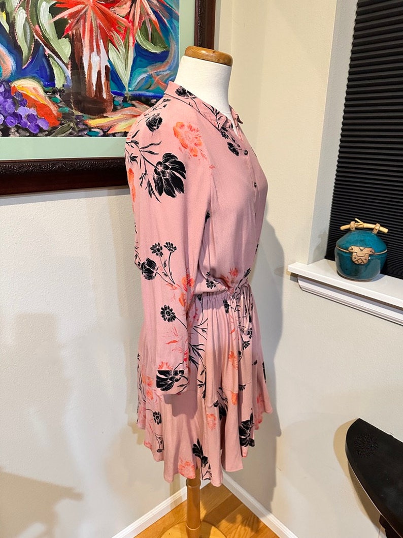 May include: A pink floral dress with black and orange floral print. The dress has long sleeves and a button-up front. The dress has a cinched waist and a ruffled hem.