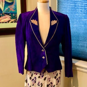May include: A vibrant purple blazer with a tailored fit, featuring a cream-colored trim and decorative leaf accents on the lapels. The blazer has two buttons and is paired with a floral print skirt, creating a vintage aesthetic.