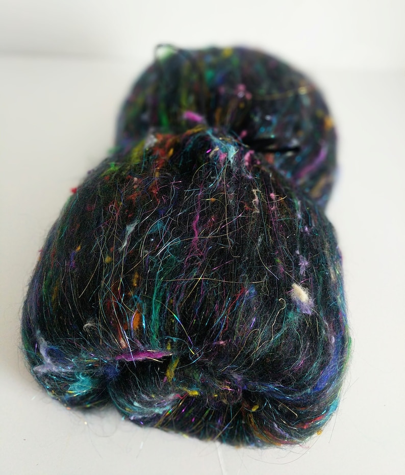 May include: A black yarn with colorful fibers and iridescent threads. The yarn is fluffy and textured.