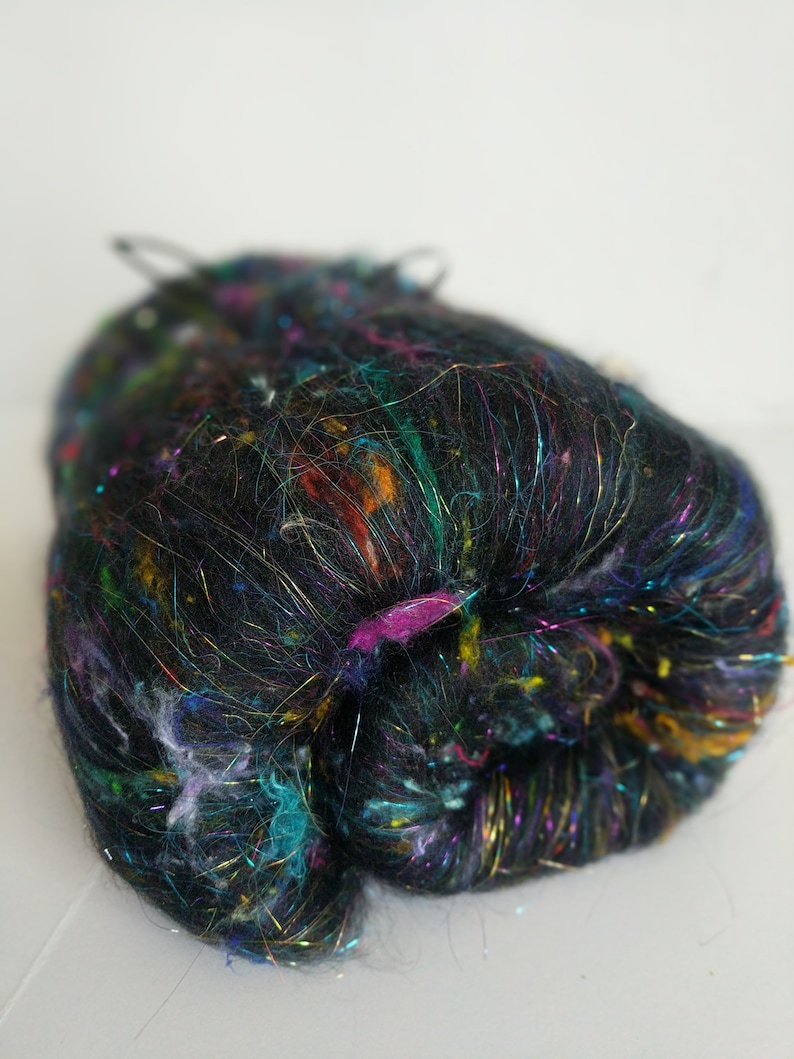 May include: A ball of black yarn with iridescent and colorful fibers. The yarn is made of a blend of fibers, including silk, wool, and metallic threads.