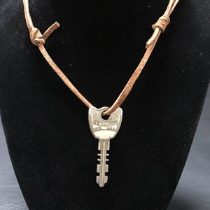 May include: A silver key pendant necklace with a brown leather cord. The key is stamped with "MILWAUKEE WIS USA". The necklace is displayed on a black mannequin.