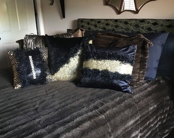 May include: A bed with a brown faux fur throw blanket and several decorative pillows. The pillows are black with gold accents and have a fluffy texture.