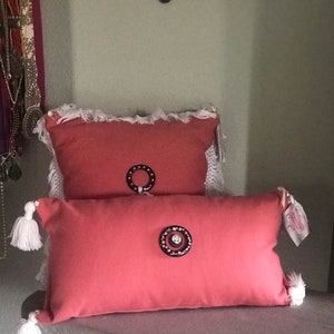 May include: Two decorative throw pillows with a dusty rose color. Both pillows have a white fringe trim and a large, round button in the center. The button on the smaller pillow is a simple, dark brown circle. The button on the larger pillow is a dark brown circle with a clear, sparkly center.