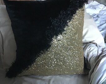 May include: A black and gold decorative pillow with a fringe trim. The pillow is made of a soft, plush fabric and has a sparkly gold finish.