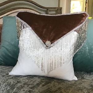 May include: A white pillow with a brown velvet accent and silver beaded fringe. The pillow has a decorative silver button in the center.