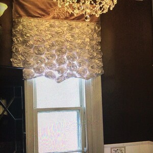 May include: White fabric window valance with a ruffled design resembling a cluster of roses. The valance is mounted above a window with white trim.