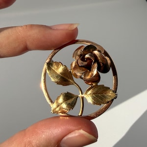 May include: A circular gold-toned brooch featuring a three-dimensional rose flower at the top right and two detailed leaves extending from a central stem, all enclosed within a thin, polished gold-colored circular frame with a metallic finish.