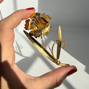 May include: A gold-toned metal flower brooch featuring a single bloom with scalloped petals and three small, round, deep blue stones set at the center. The design includes a long, slender stem and two pointed leaves, one of which has a curled tip.