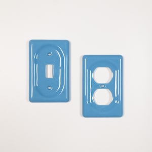 May include: Two light blue electrical wall plates. One plate has a rectangular opening for a light switch. The other plate has two circular openings for an electrical outlet. Both plates are rectangular with rounded corners.