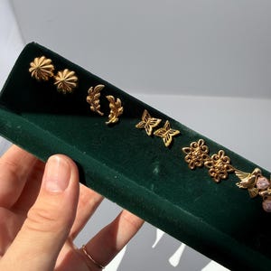 May include: A collection of gold-colored stud earrings displayed on a dark green velvet surface. The earrings feature various designs, including floral, leaf, and butterfly shapes. The earrings are presented in a row, showcasing their intricate details and craftsmanship.