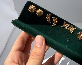May include: A collection of gold-colored stud earrings displayed on a dark green velvet tray. The earrings feature various designs, including flower, leaf, butterfly, and floral patterns. The earrings are presented in a row, showcasing their different shapes and styles.