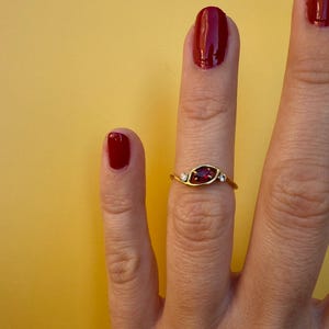 May include: A gold ring featuring a central oval-shaped red gemstone, flanked by two smaller white gemstones. The ring is worn on a finger with red nail polish against a yellow background.