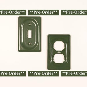 May include: Two rectangular olive green wall plates with a glossy finish. One plate features a single vertical toggle switch opening, while the other has two circular outlet openings. Each plate includes the text **Pre-Order**.