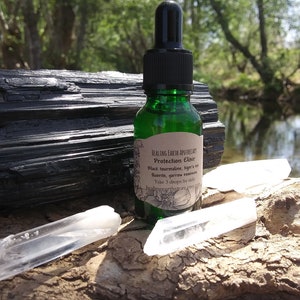 May include: A green glass bottle with a dropper filled with a clear liquid. The bottle has a label that reads "Healing Earth Apothecary Protection Elixir Black Tourmaline, Tiger's Eye, Fluorite, Yarrow essential oil Take 3 drops for daily healing. healingearthstore.com". The bottle is sitting on a piece of wood with a black tourmaline crystal and two clear quartz crystals.