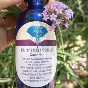 May include: A blue glass bottle with a white label that reads "HEALING EARTH APOTHECARY Ganesha Om Gum Ganapataye Namah Spray to remove obstacles and invoke Ganesha Sandalwood, Elemi, Chamomile essential oils, vodka, Reiki infused Sedona Spring water, love, 432 hz healingearthstore.etsy.com". The bottle is decorated with a blue elephant graphic.