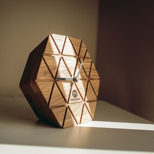 Pode incluir: Um relógio de madeira com um design geométrico e um mostrador branco. O relógio é feito de madeira marrom claro e tem um padrão triangular. O relógio está sobre uma superfície branca.