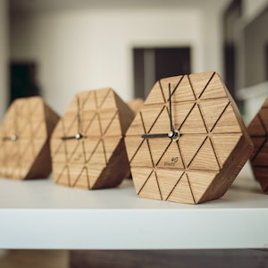 Könnte beinhalten: Vier sechseckige Holzuhren mit schwarzen Zeigern und einem geometrischen Design. Die Uhren sind in einer Reihe auf einer weißen Oberfläche angeordnet. Jede Uhr hat das Wort "Enretic" auf dem Zifferblatt.