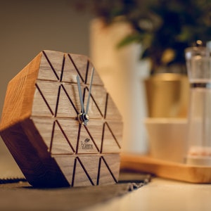 May include: A hexagonal wooden clock with black hands and a geometric design. The clock is made of light brown wood and has a small inscription on the face that reads "Wooden Wonders".