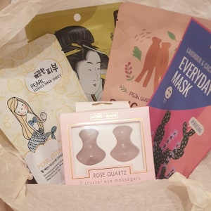 May include: A pink box containing a selection of beauty products. The box includes two face masks, a pair of rose quartz eye massagers, and a sheet mask. The face masks are labelled 'Everyday Mask' and 'Pearl Essence Mask Sheet'. The eye massagers are labelled 'Rose Quartz 2 crystal eye massagers'.