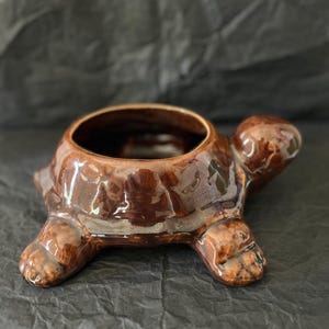 May include: A brown glazed ceramic planter shaped like a turtle. The turtle figure features a glossy finish with mottled dark and light brown tones, a rounded shell opening at the top, a protruding head, and four short legs extending from the base.